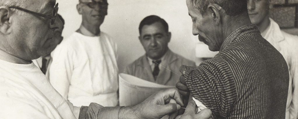 A imagem mostra cinco homens, sendo três médicos ou enfermeiros, um migrante e um que aparenta ser colaborador da Hospedaria, por estar de terno e gravata. O migrante está tomando vacina.