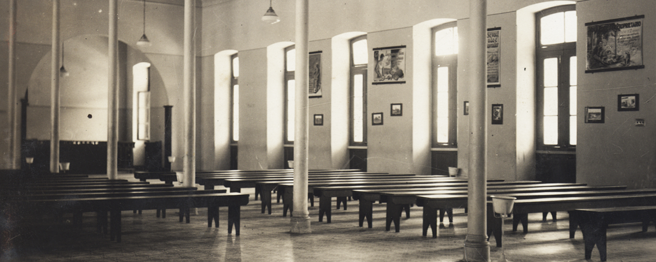 A imagem mostra, em uma sala, bancos compridos dispostos em duas filas, com pilastras no meio