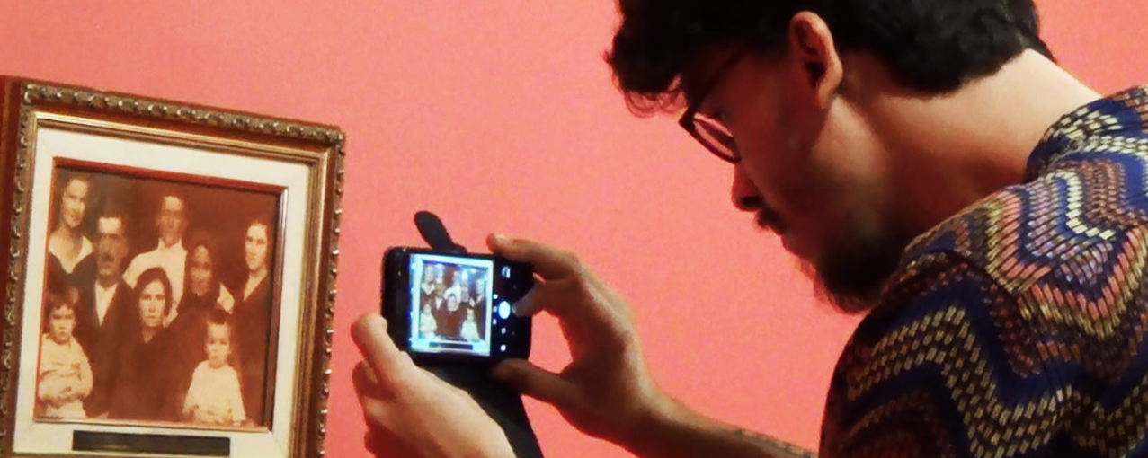 Um retrato de família está pendurado em um quadro na parede e um jovem está em frente tirando foto dessa imagem com o celular