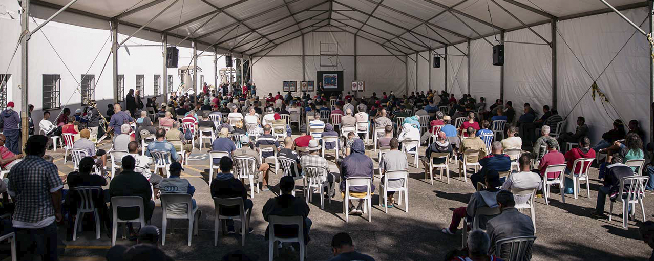 A imagem mostra, de costas, muitos homens sentados em cadeiras brancas, embaixo de uma tenda grande da mesma cor