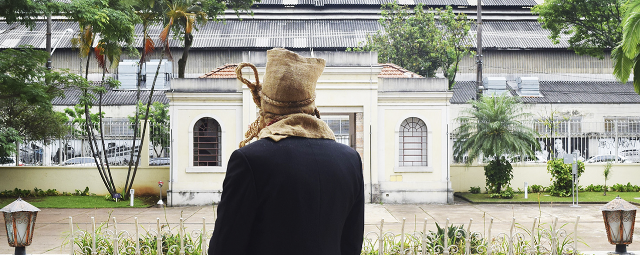 Fotografia retangular, colorida, mostrando, de costas, um homem com a cabeça tampada por junta. Na frente dele, aparece parte do jardim do Museu da Imigração