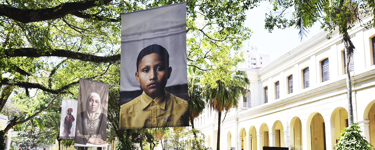 A imagem mostra fotos em grande escala dispostas no jardim do Museu da Imigração, penduradas nas árvores