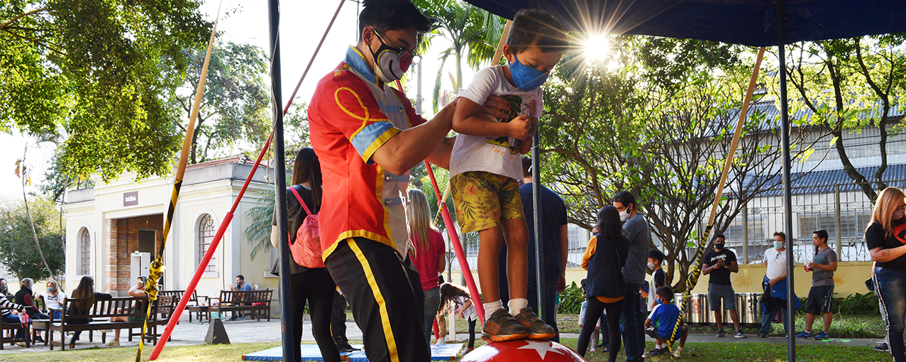 Imagem de uma oficina de circo com um profissional segurando um menino em cima de uma bola
